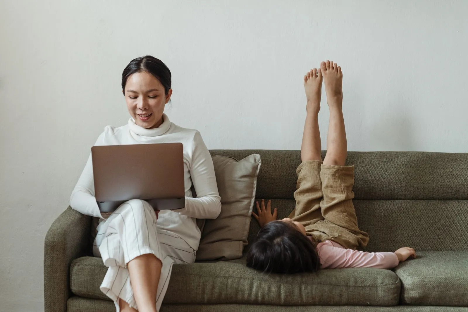 A parent working on a laptop at home while a child plays nearby on the couch